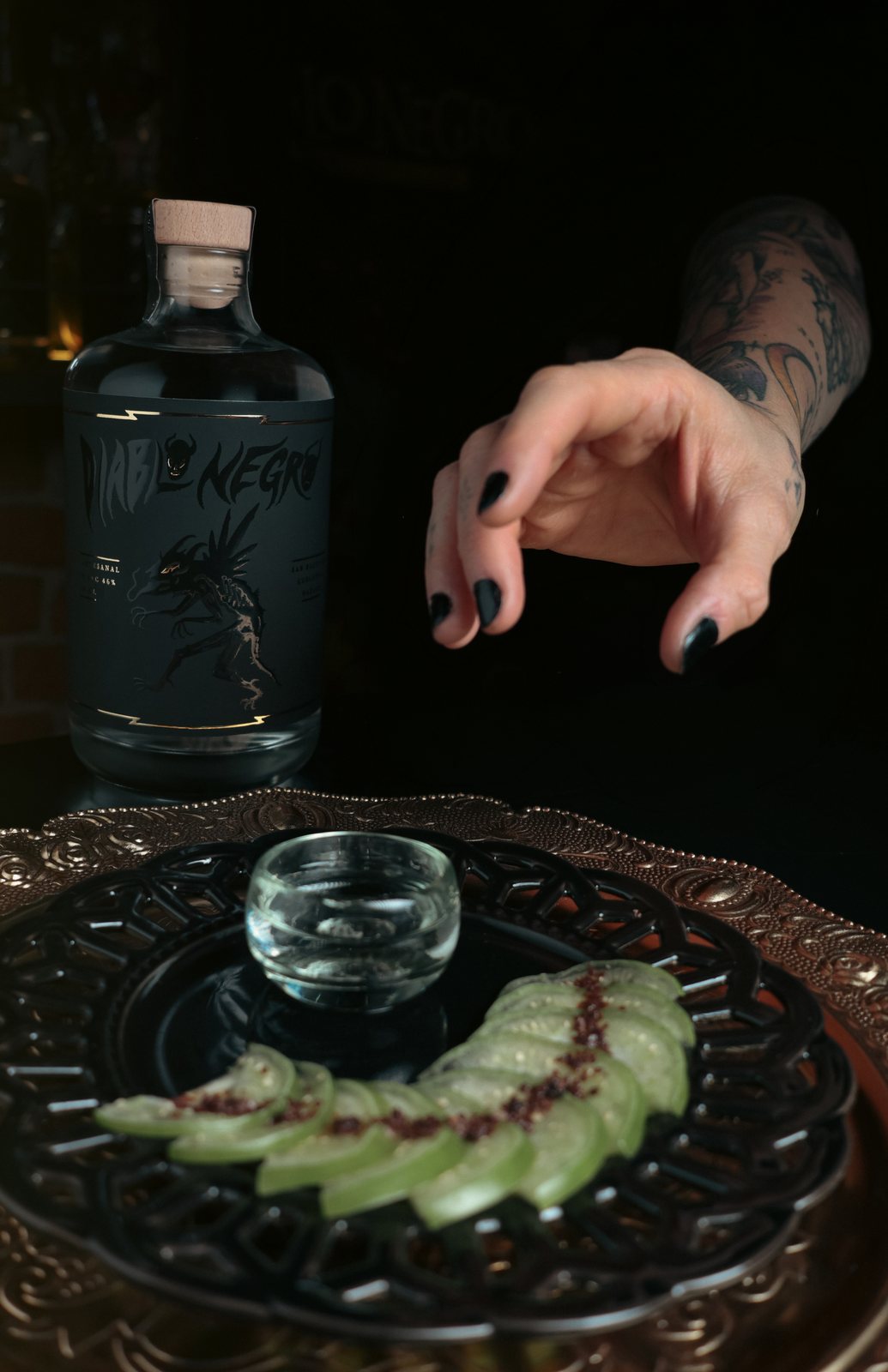Una mano con uñas negras se acerca a una copita de mezcal servida junto a la botella de Diablo Negro y rodajas verdes con sal