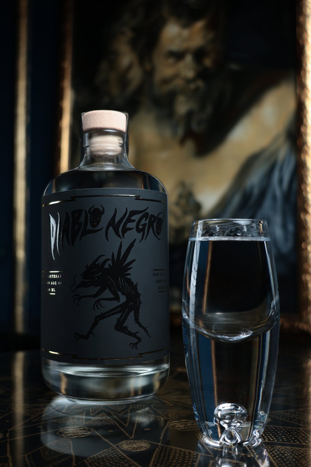 Botella de Mezcal Diablo Negro junto a un caballito servido, frente a una pintura antigua